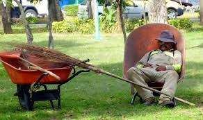 Mies nukkuu vanhassa tuolissa nurmikolla hattu silmillä. Vieressä on kottikärryt, joihin nojaa luuta.