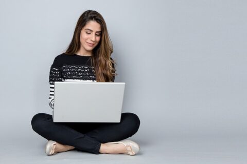 Nainen istuu lattialla jalat ristissä ja kirjoittaa kannettavalla tietokoneella.