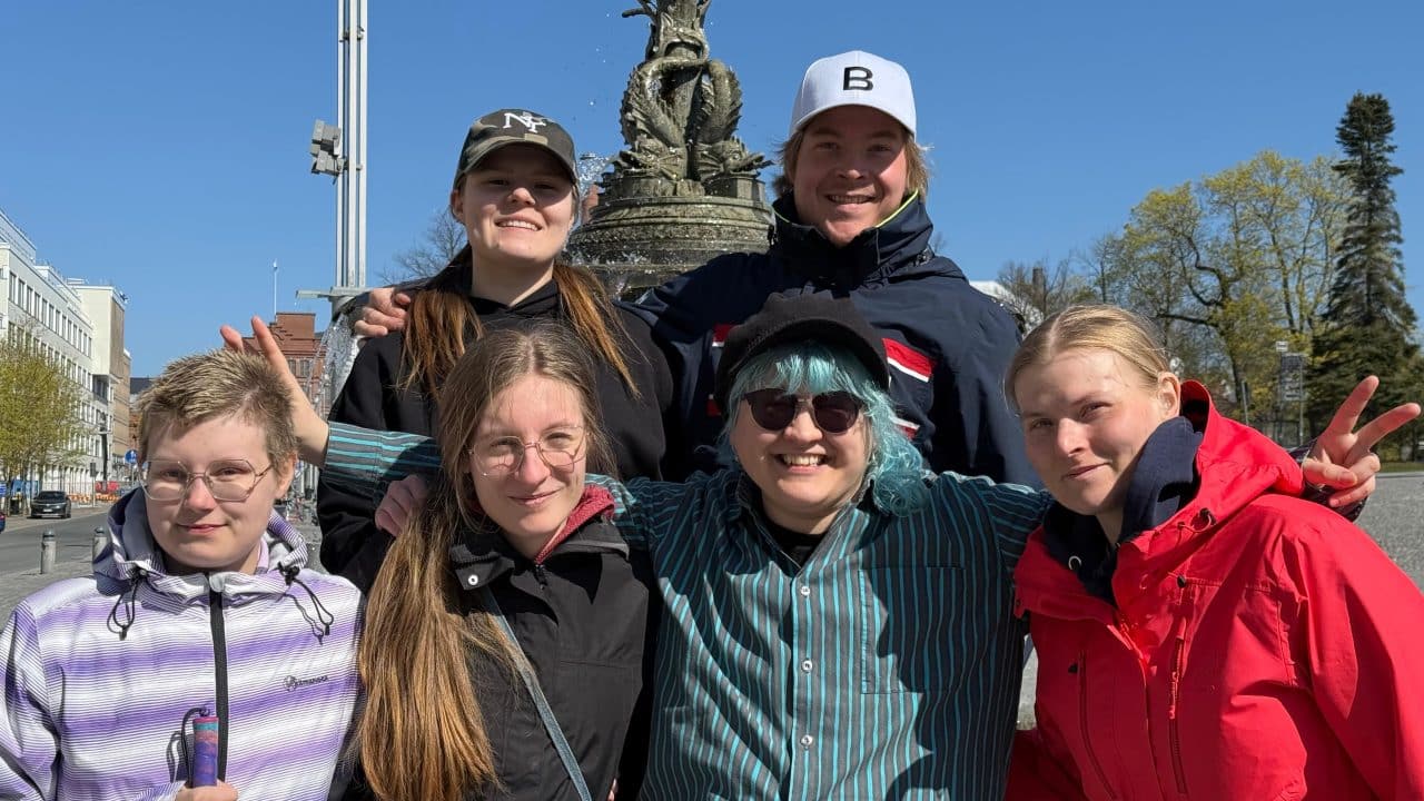 Kuusi nuorta aikuista poseeraa iloisina kirkkaassa kevätauringossa suihkulähteen edessä. He hymyilevät ja seisovat lähekkäin, osa tekee rauhanmerkkejä. Taustalla sininen taivas ja kevätpuut.