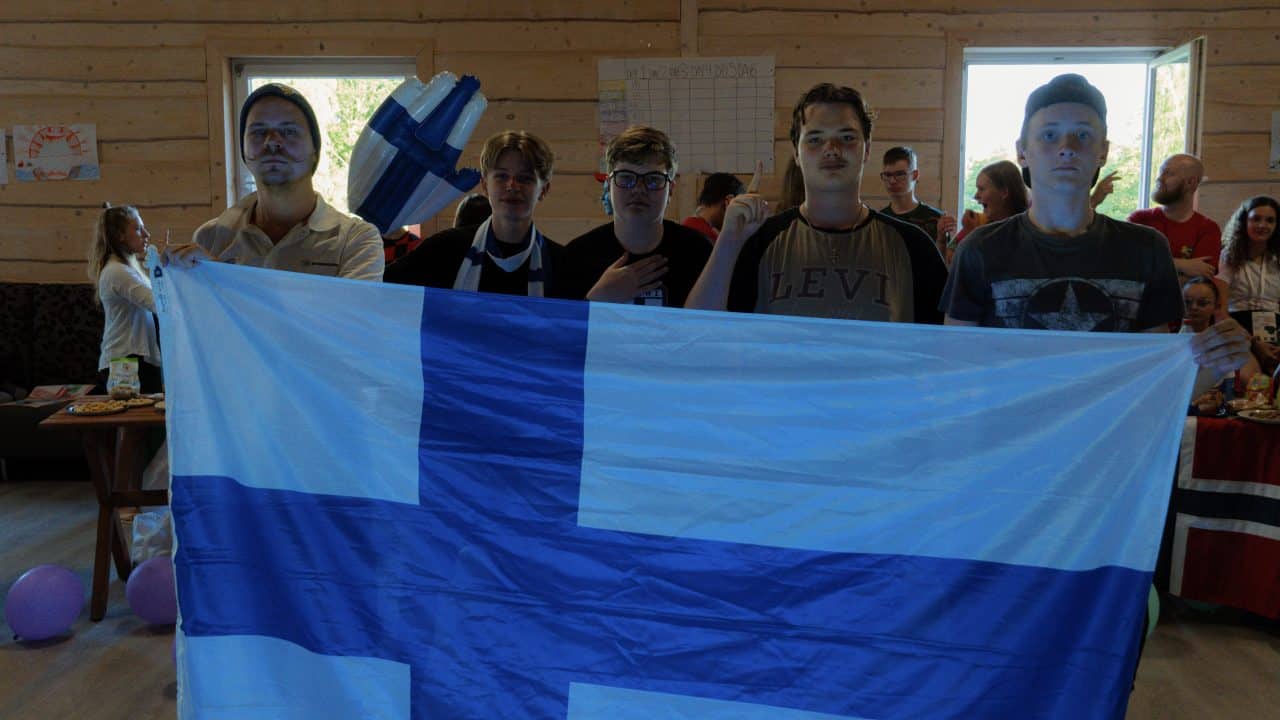 Kuvassa ohjaajan lisäksi 4 nuorta seisoo sisätilassa pitäen edessään suurta Suomen lippua. He katsovat vakavina (suomalaisina) kohti kameraa. Taustalla on muita ihmisiä, koristeita ja pöytiä, joiden päällä on ruokaa ja juomia. Yhdellä pöydällä näkyy myös Norjan lippu.