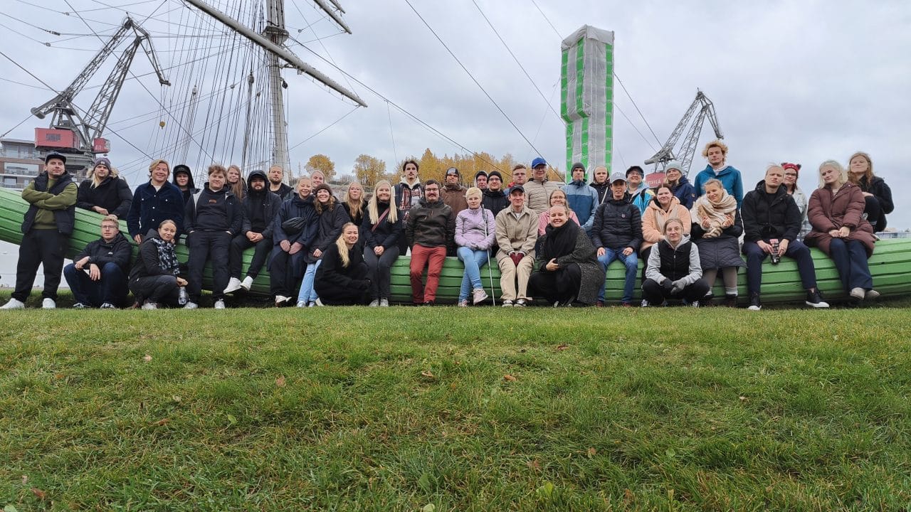 Kuvassa on suuri ryhmä ihmisiä (noin 40 henkeä), jotka poseeraavat ulkona vihreällä ruohikolla. He istuvat tai seisovat vihreän, kaarevan puurakennelman päällä, joka muistuttaa veneen kylkeä. Taustalla näkyy suuri purjelaiva, nostureita ja rakennustyömaa, jossa on vihreä-valkoinen suojapeite tornimaisessa rakenteessa. Ihmiset ovat pukeutuneet lämpimästi – takkeihin, pipoihin ja huppareihin – ja sää näyttää syksyiseltä, pilviseltä ja hieman harmaalta. Tunnelma vaikuttaa rennolta ja yhteisölliseltä.