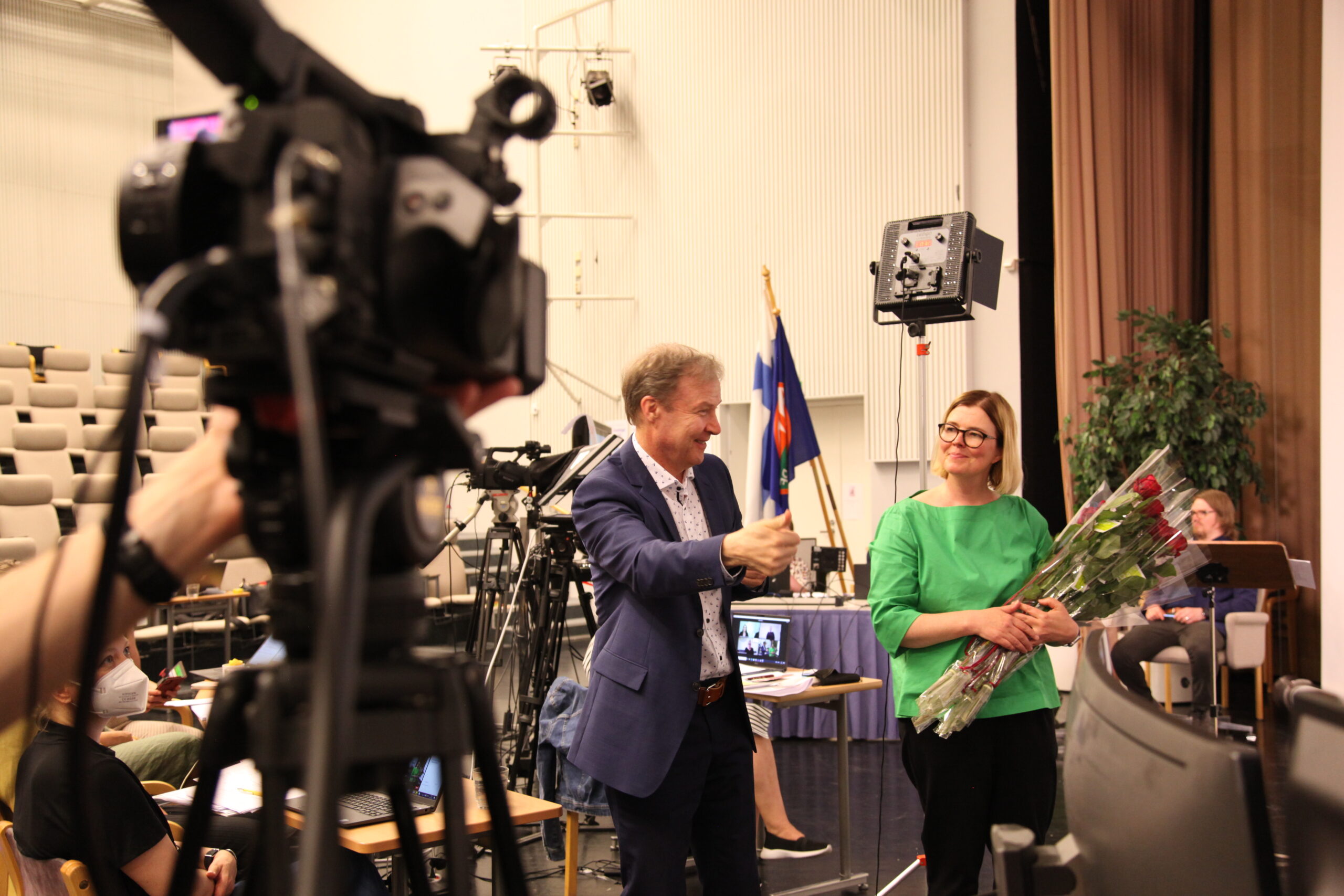 Markku Jokinen viittoo kiitoksia ja Kaisa Alanne pitää kädessää ruusunippua. Kuvan vasemmassa reunassa on kamera.
